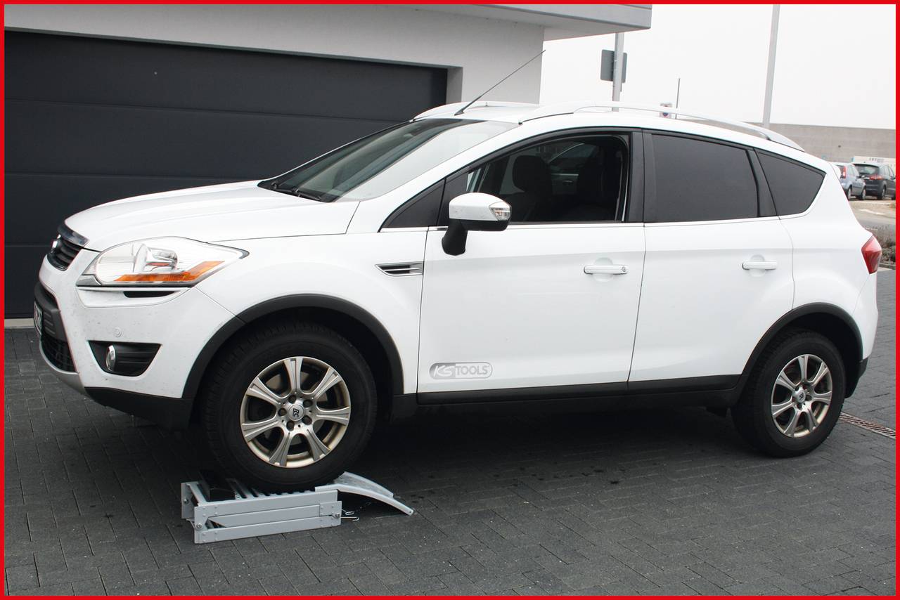 A white SUV is parked on a driveway in front of a garage. The right front wheel is resting on a ramp.