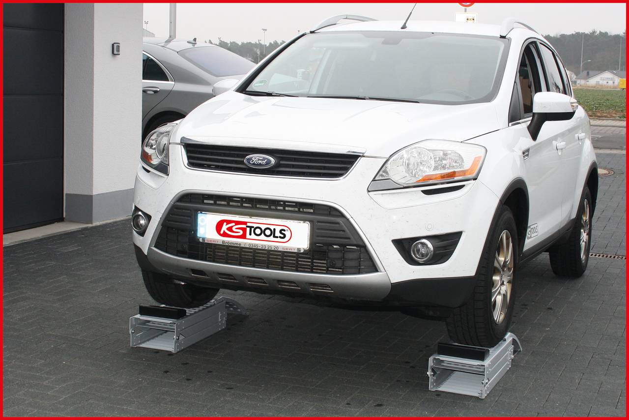 A white car is raised on two ramps from KS Tools, positioned for repair work. Another vehicle is visible in the background.