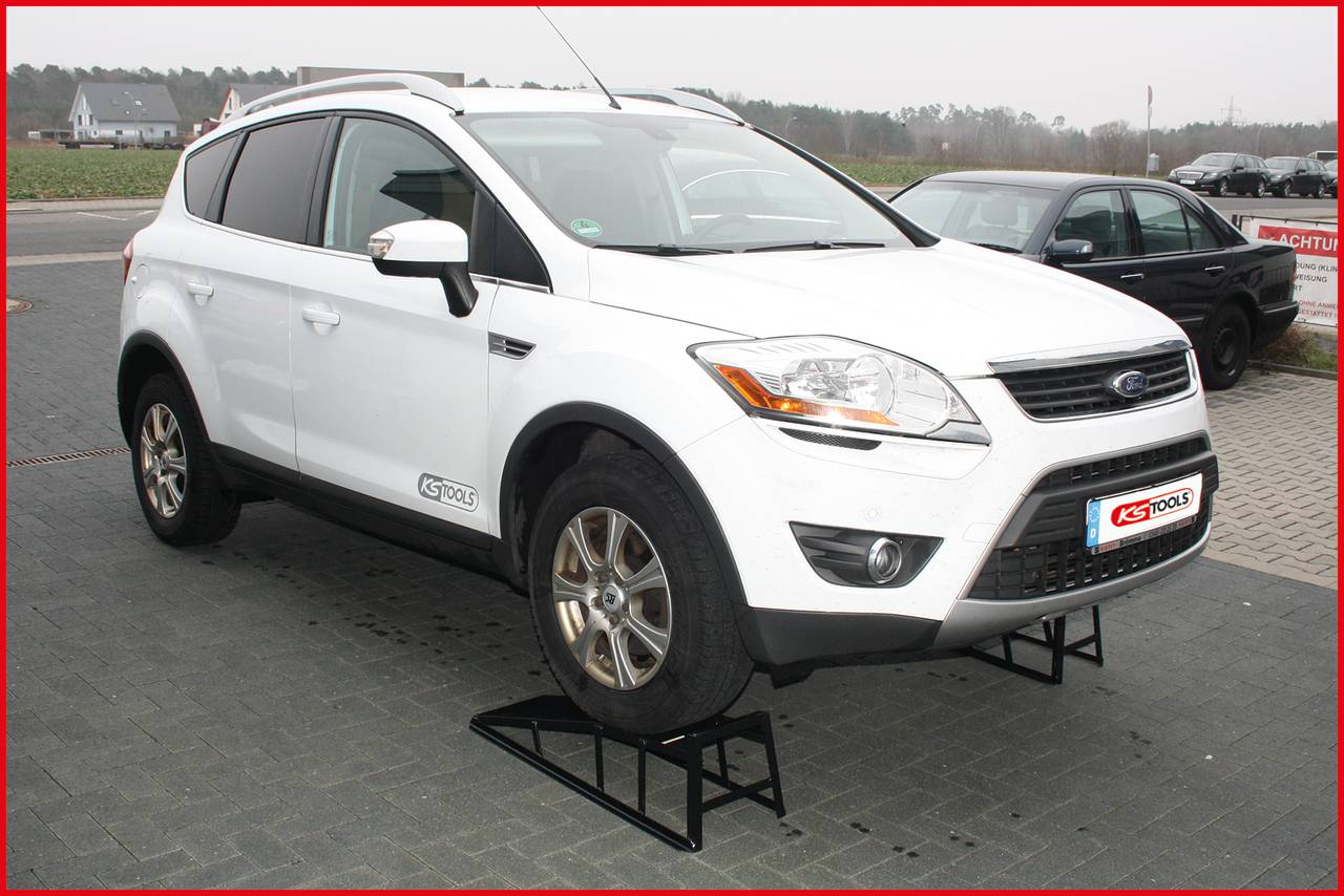A white car is raised on metal stands in an open parking area. Another car is visible in the background.