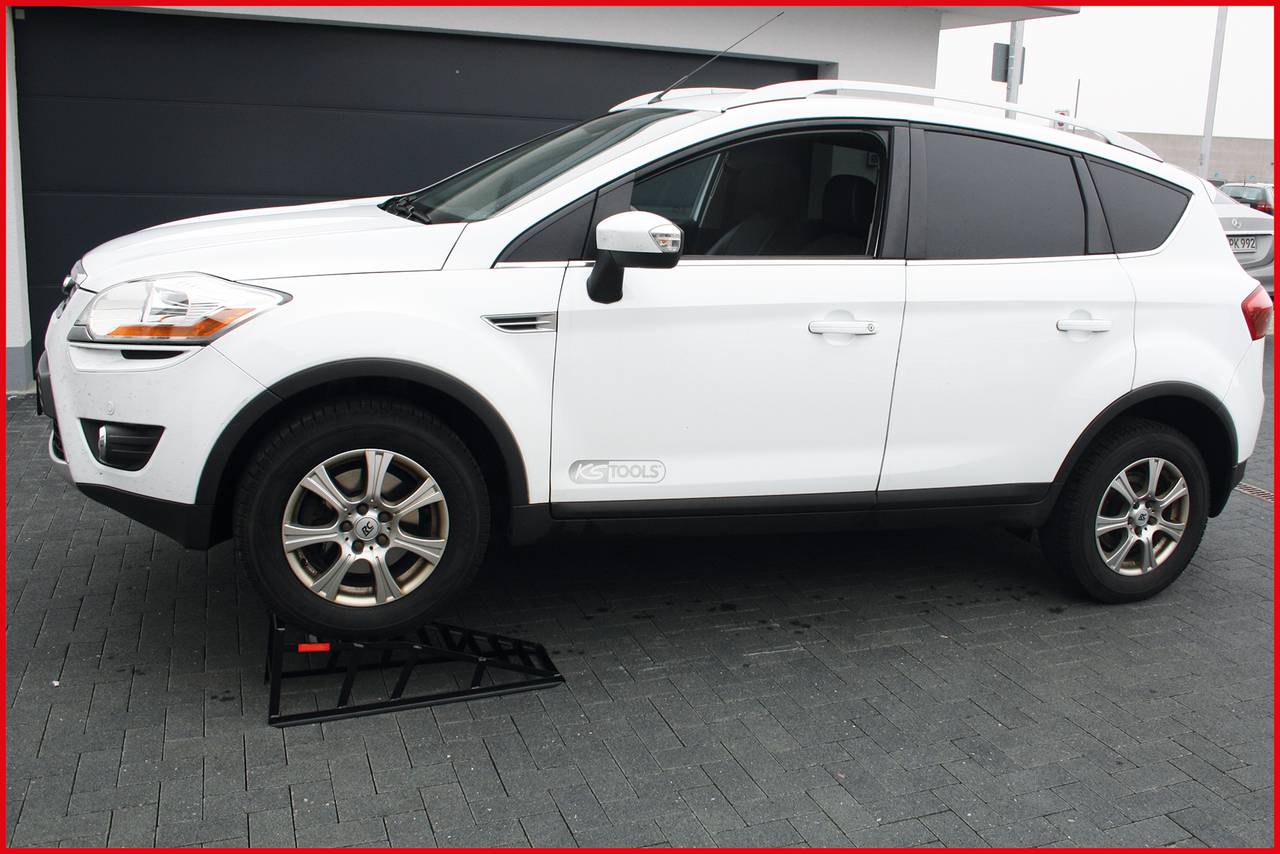 A white SUV vehicle is parked at an angle on a ramp on an asphalted surface in front of a garage.