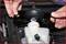 Two hands remove the lid of a brake fluid reservoir in a car's engine compartment. The container is partially filled.