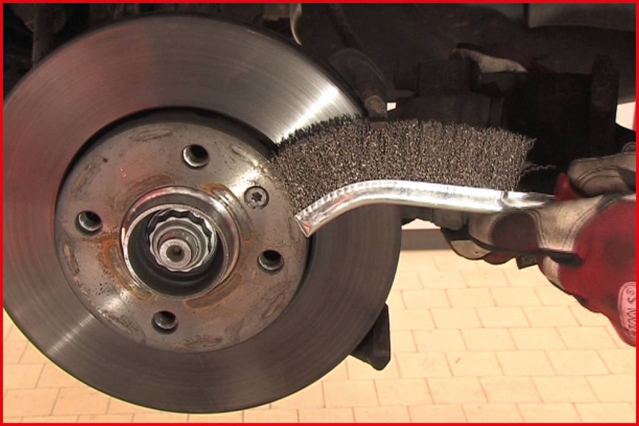 A hand is cleaning a car brake disc with a wire brush. The brake disc is firmly mounted on a vehicle jack.