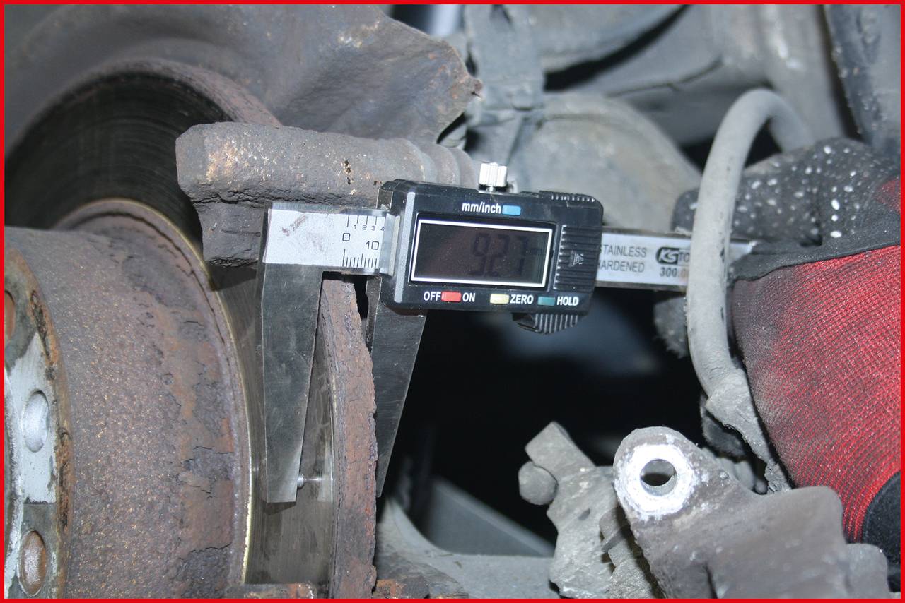 A vernier caliper measures the thickness of a brake disc, the display shows 9.27 mm. The disc is part of a car braking system.