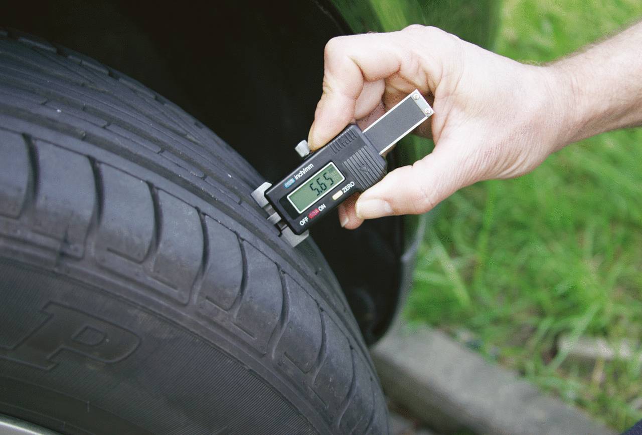 A person is measuring the tread depth of a car tyre with a digital measuring device. The screen displays '5.65'.