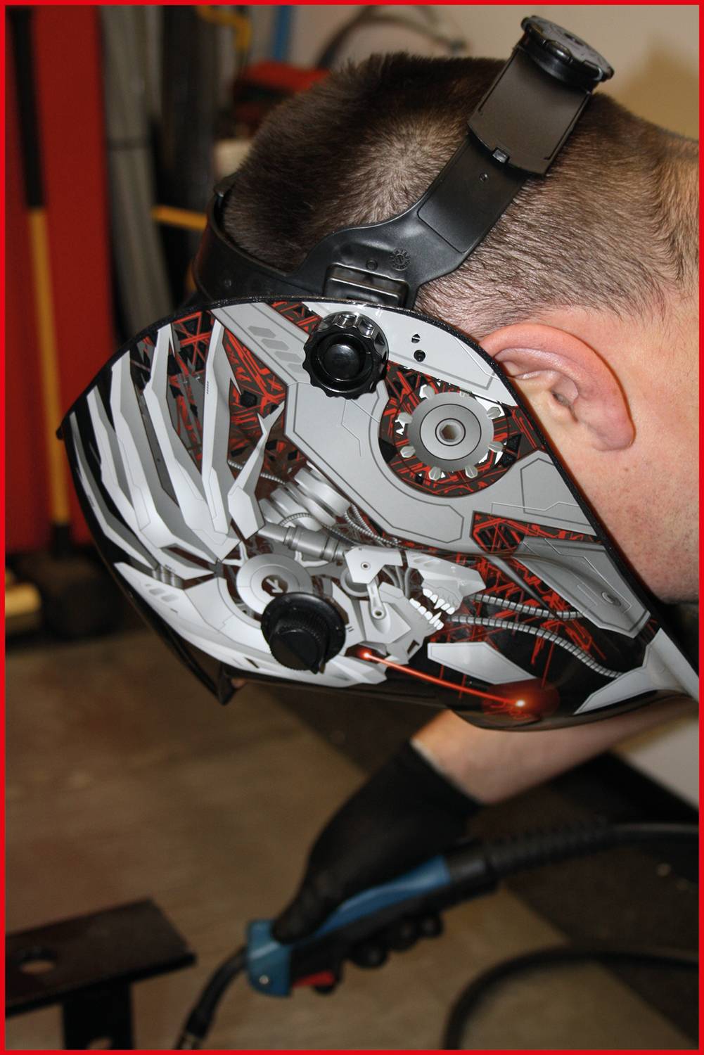 A person is wearing a welding helmet with a modern design and holding a welding device. The environment suggests a workshop.