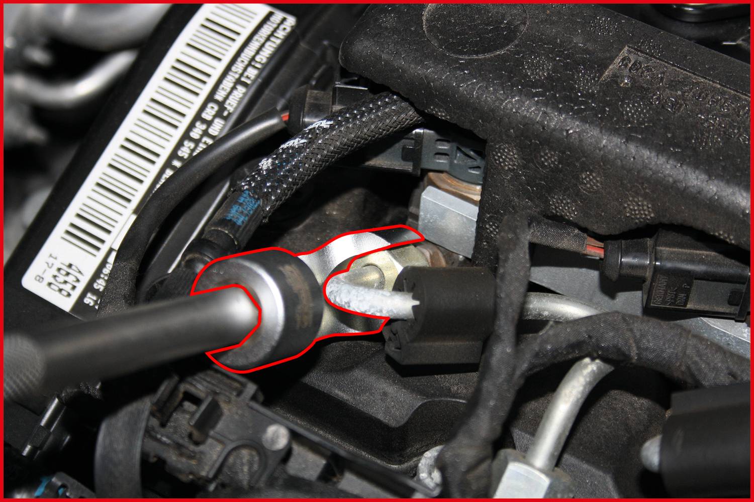 Close-up of a car engine focusing on a metal connector being held by a tool. The surrounding environment shows cables and hoses.