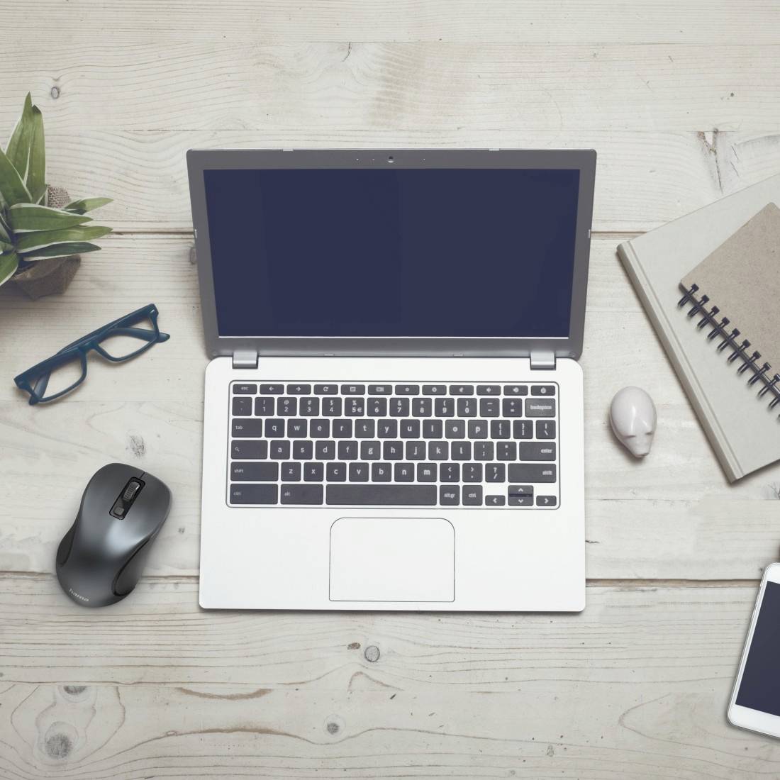 A laptop on a wooden table, surrounded by a pair of glasses, a notebook, a mouse and a plant.