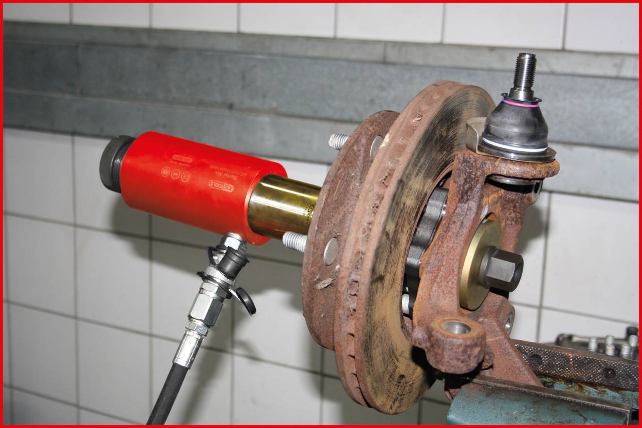 An automotive part is being machined with a red pneumatic tool. The workpiece is rusty and is located in a workshop environment.