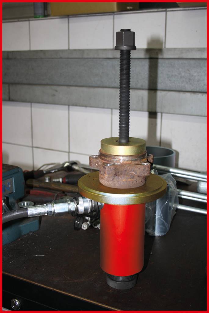 A red hydraulic tool sits on a workbench in a workshop. It is surrounded by various tools and accessories.