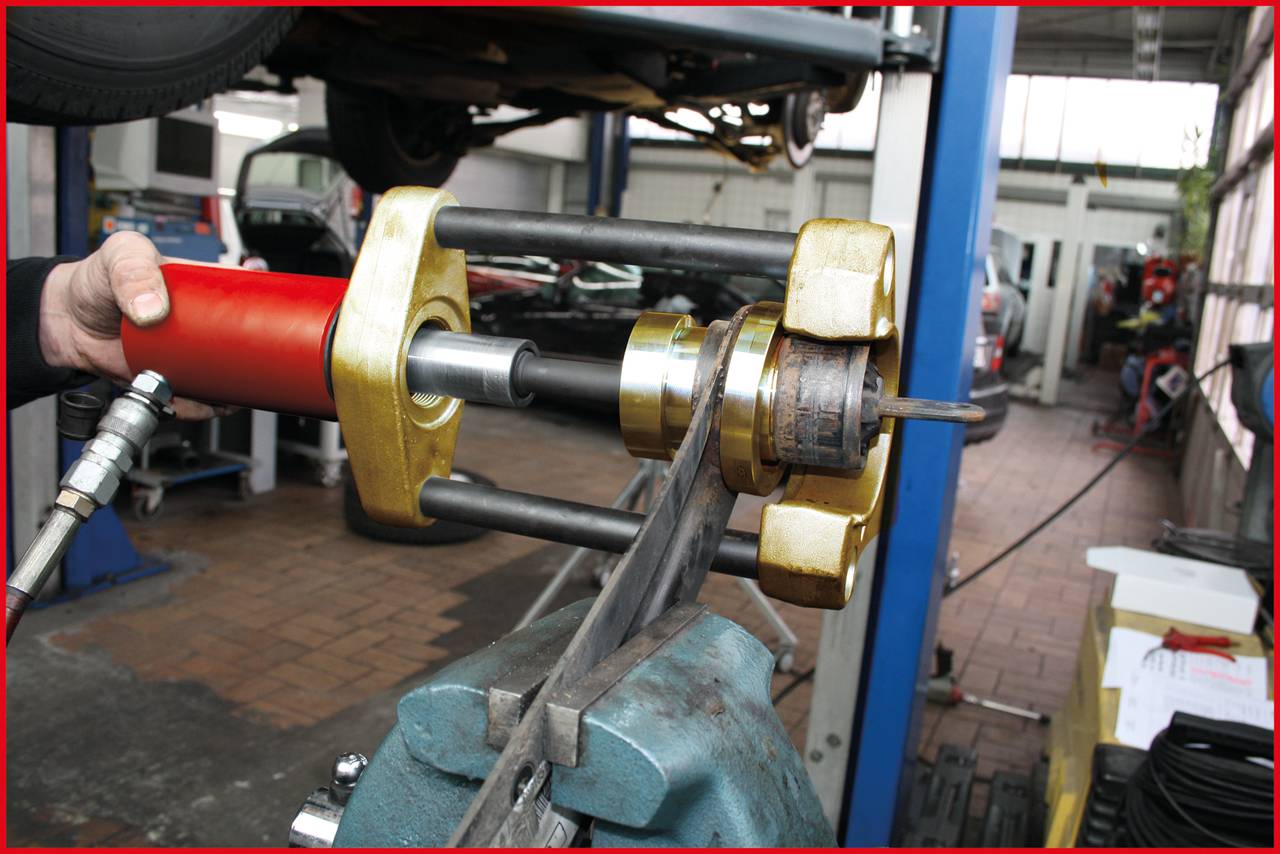 A mechanic is using a specialised tool in a workshop to repair or assemble a metal part. Cars are visible in the background.
