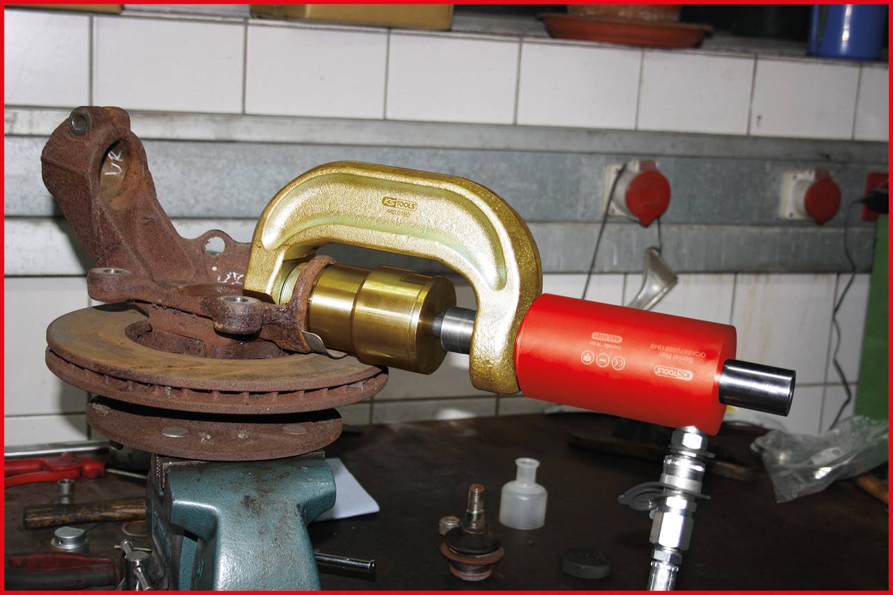 A hydraulic tool deforms a metal shaft that is clamped in a vice. Tools can be seen in the background.