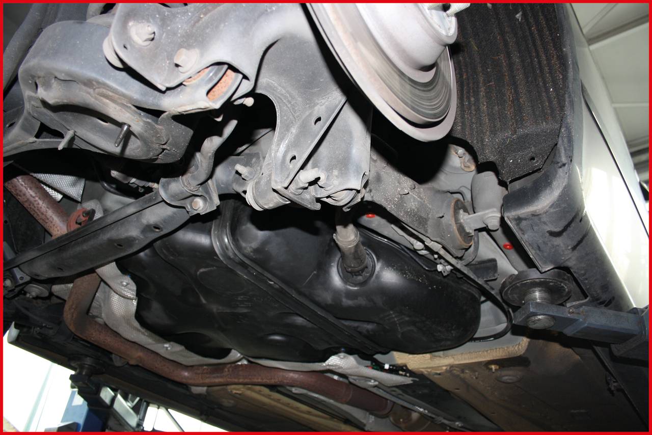 Underside of a vehicle in a workshop. View of exhaust system, fuel tank and chassis components during repair inspection.