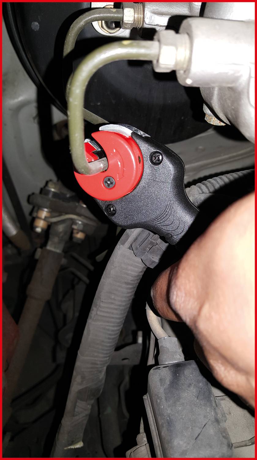 Engine compartment of a vehicle with a close-up of a component featuring a red plastic part and hoses. A hand is working on it.