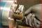 A hand in a protective glove is holding a metal tool against a cylindrical metal machine, possibly in a workshop.