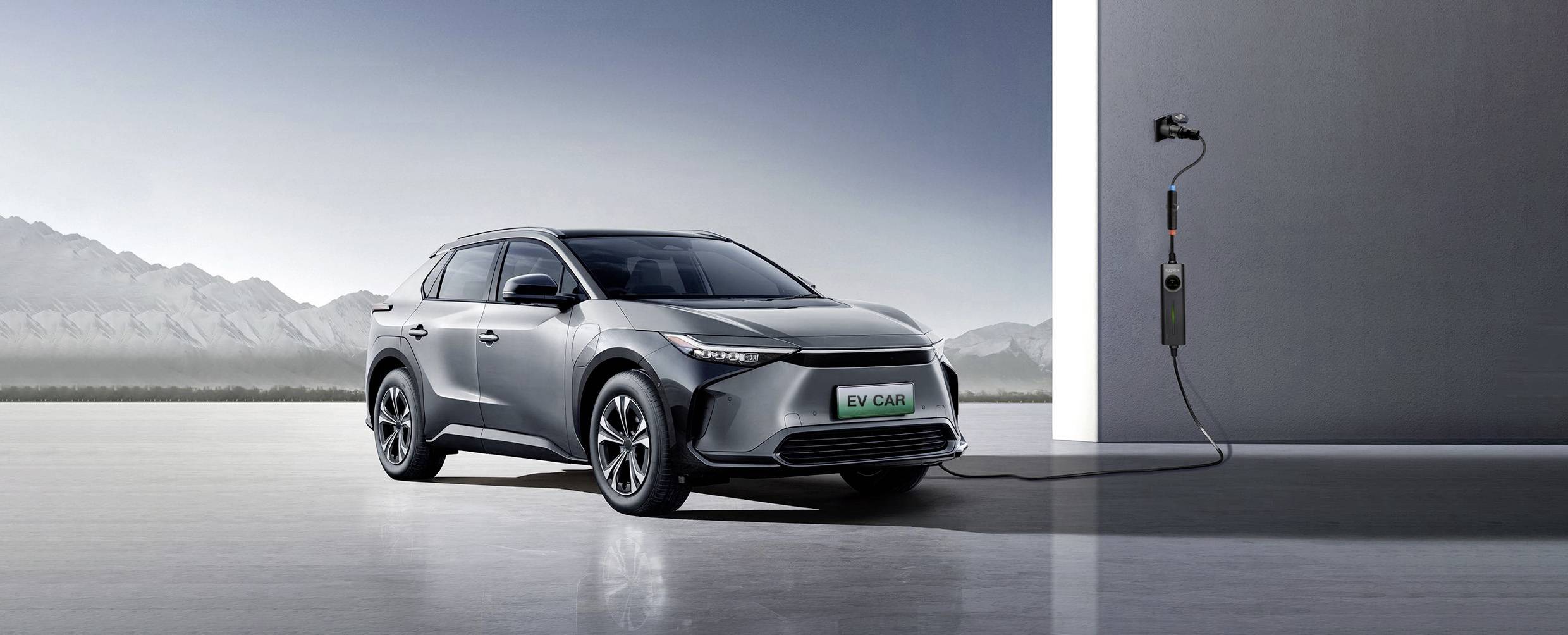 A grey electric car is charging at a charging station in a modern, minimalist environment with mountains in the background.
