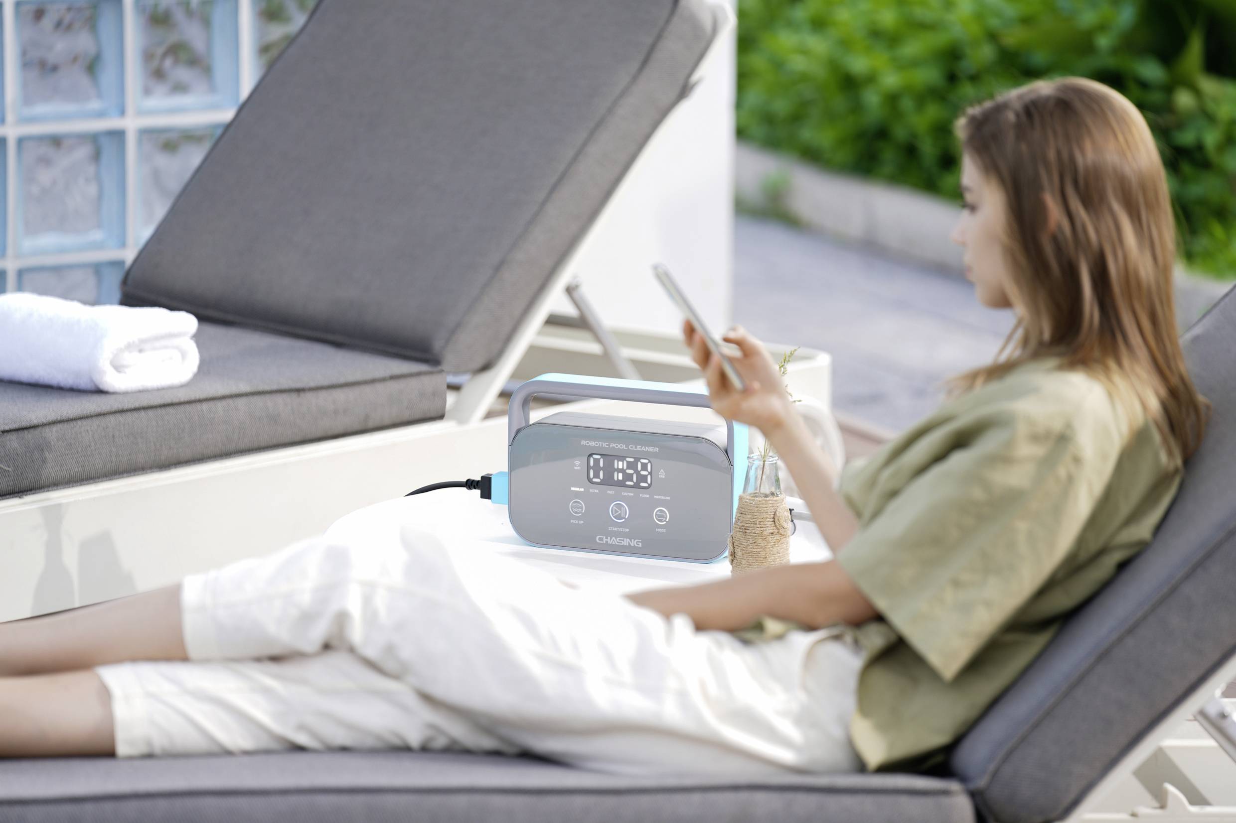 A woman is relaxing on a lounger outdoors and looking at her mobile phone, with a portable device beside her.