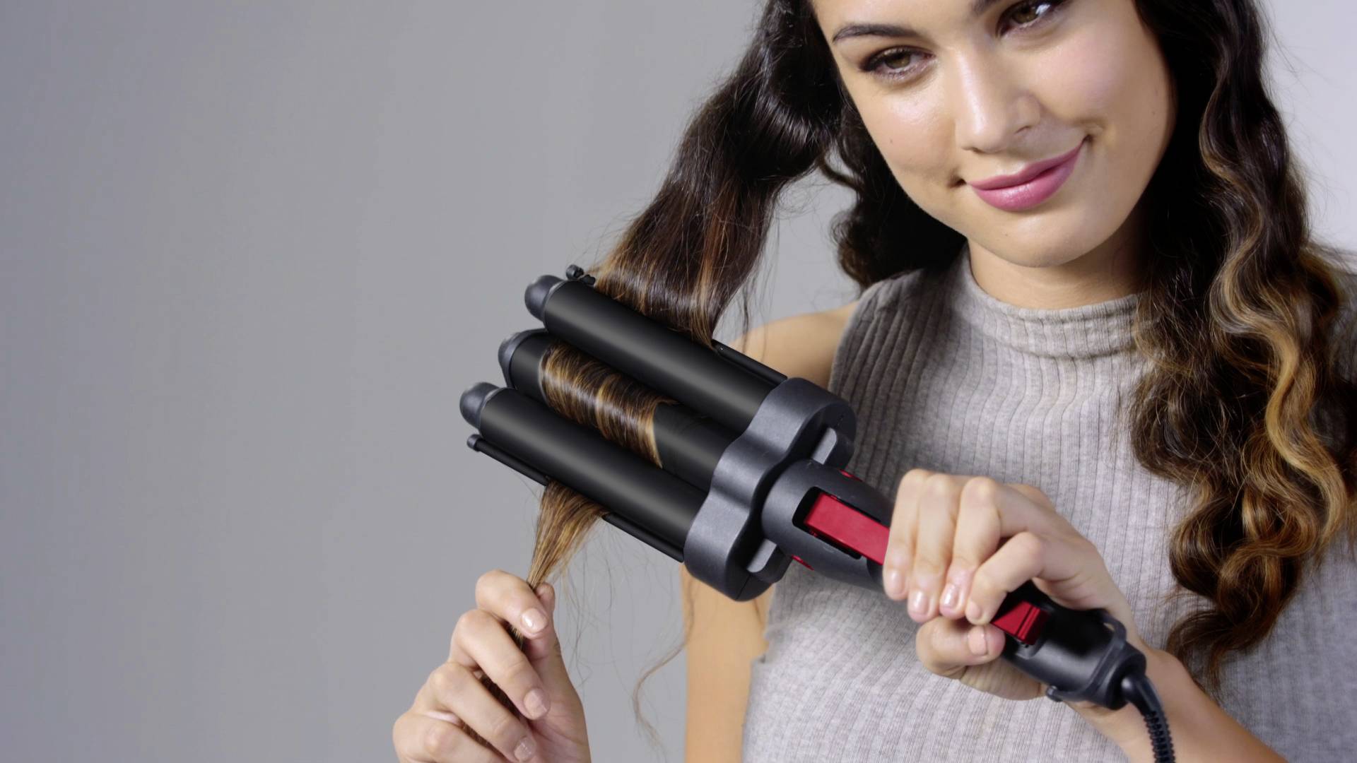 A woman is using a curling wand with three barrels to style her hair. She is wearing a sleeveless top.