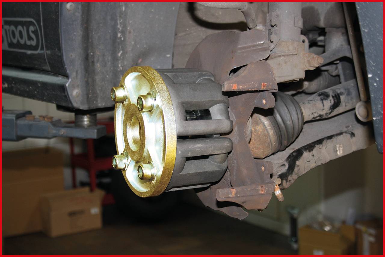 Close-up of a vehicle suspension, showing a brake component without a wheel. The surroundings suggest a workshop with tools and cardboard boxes.