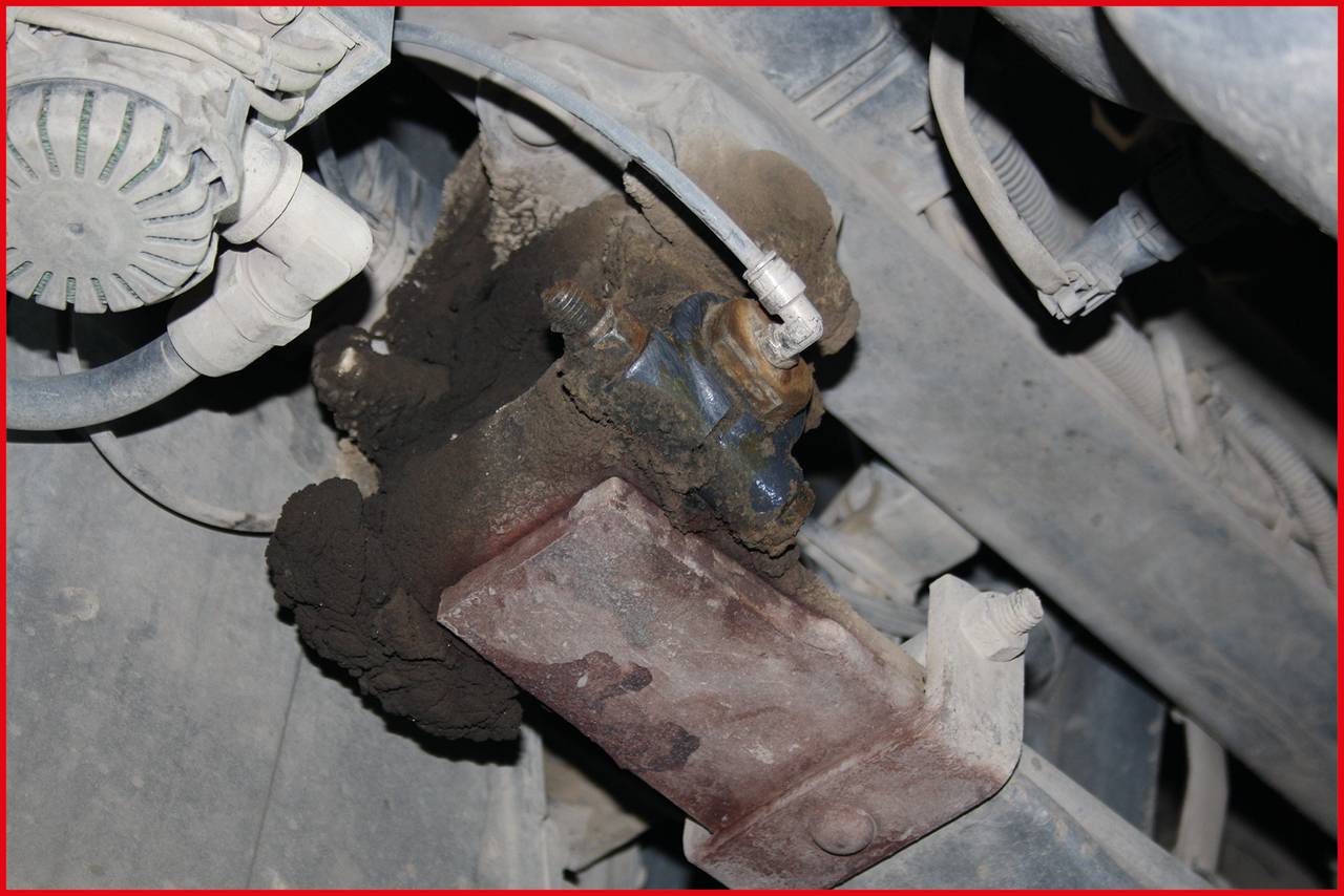 Close-up of a machine component covered in dirt in a vehicle engine bay, showing wear and dirt accumulation on bolt connections.