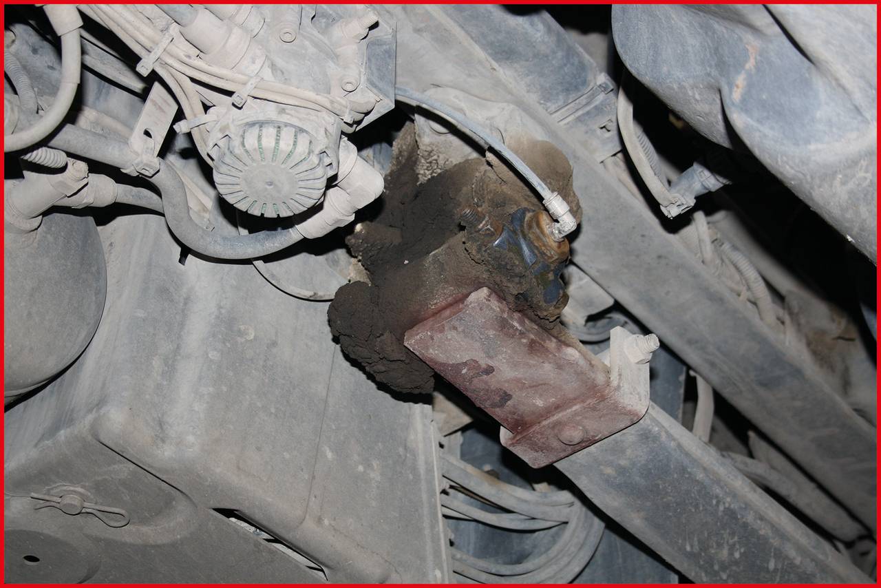 A rusty metal component beneath a vehicle chassis, surrounded by cables and pipes. It shows signs of wear and dirt accumulation.