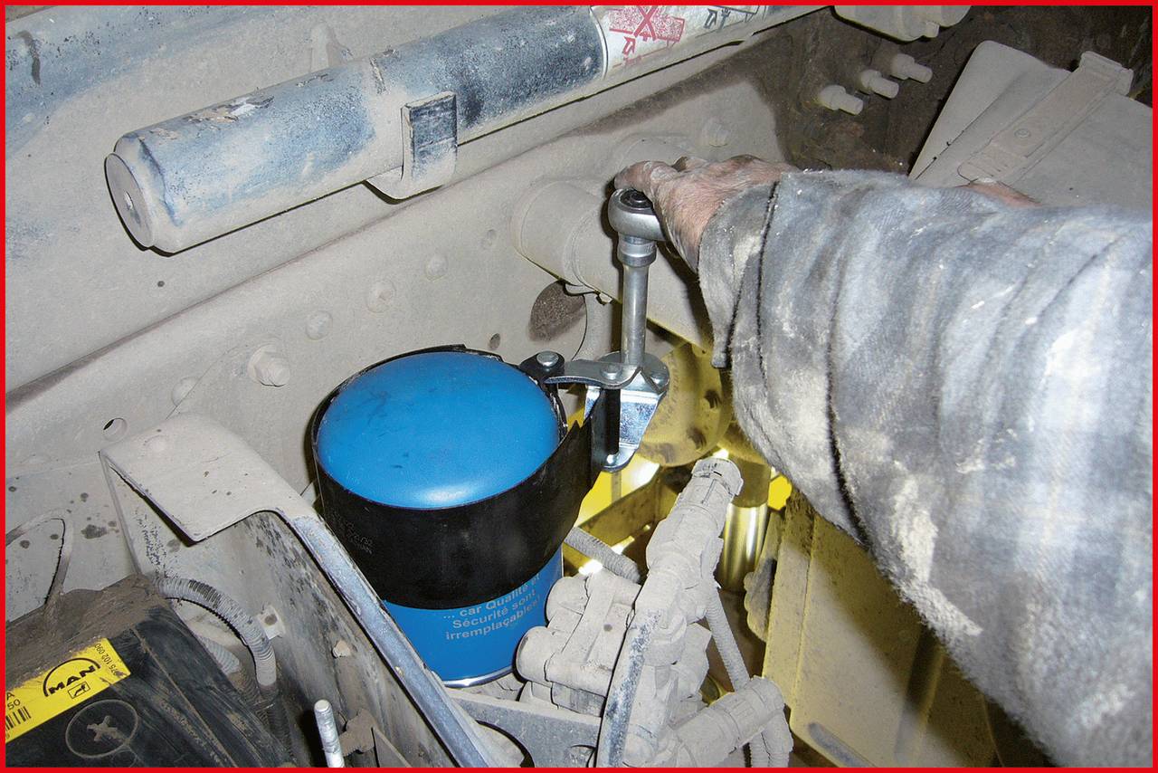 A worker is using a tool to adjust a machine. A blue filter is visible. The environment is dusty and industrial.