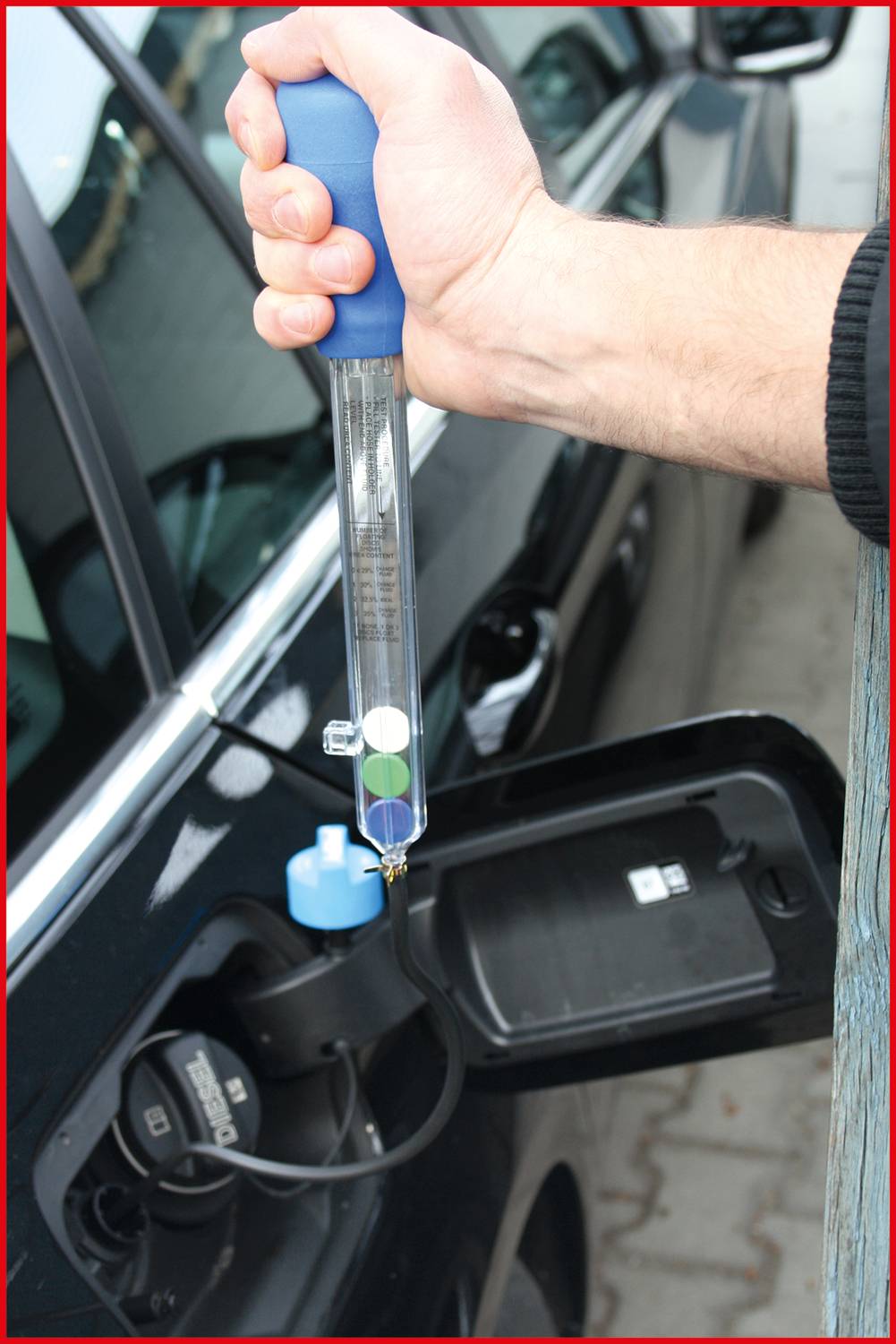 A person is checking the antifreeze in a car using a hydrometer. The hydrometer indicates different densities through coloured floats.