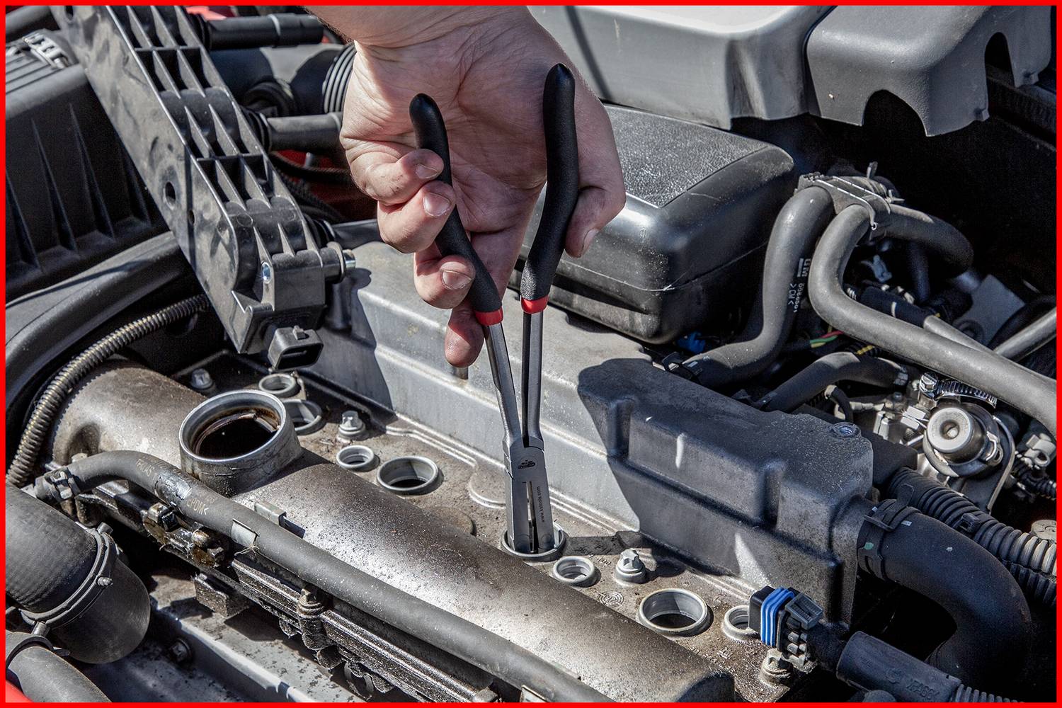A hand holding pliers is pulling a spark plug lead out of an open car engine compartment.