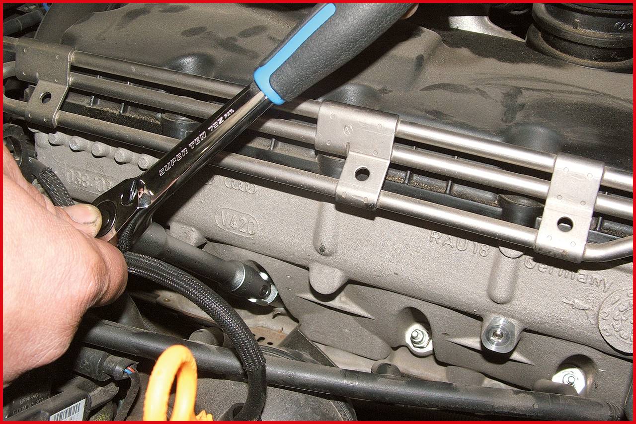 A hand is holding a spanner working on a valve cover of a car engine. The surrounding area shows various engine components.