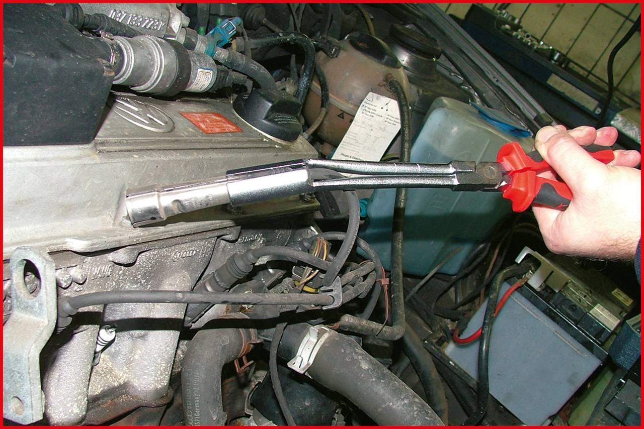A hand is holding a spark plug socket on a spanner over an open engine compartment of a car.