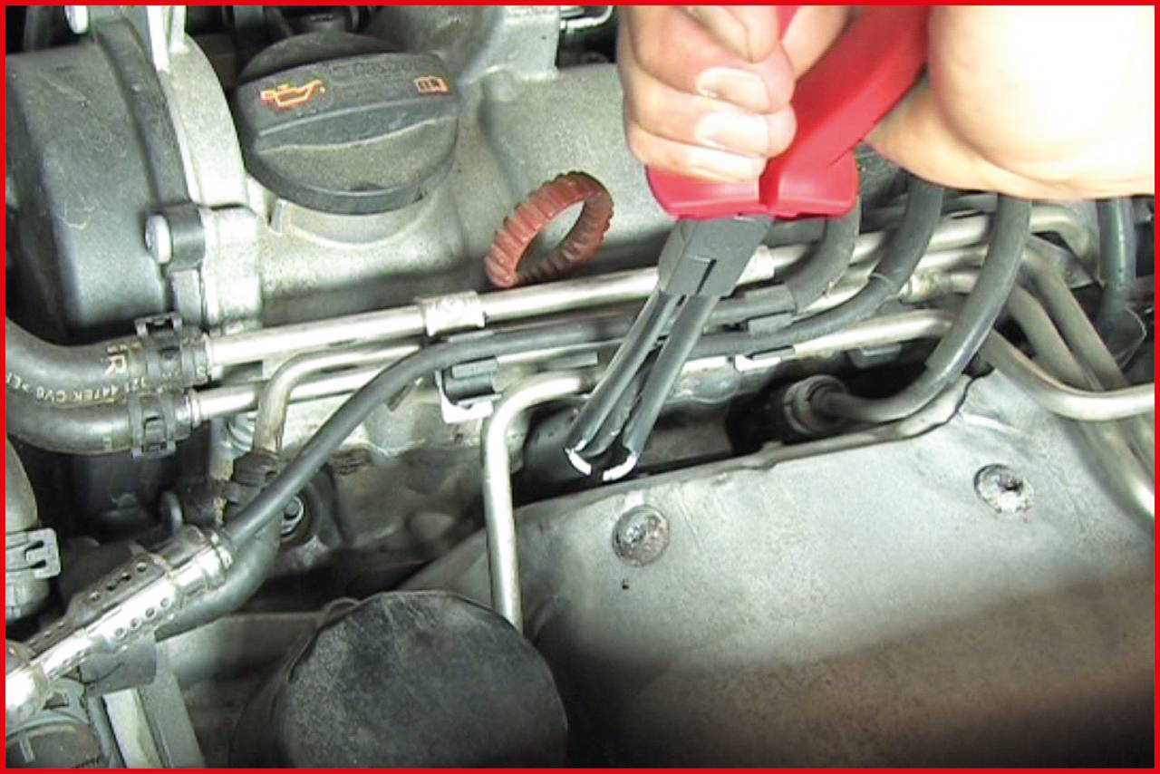 A hand is using pliers to remove an engine clip. Various engine components and several cables are visible in the background.