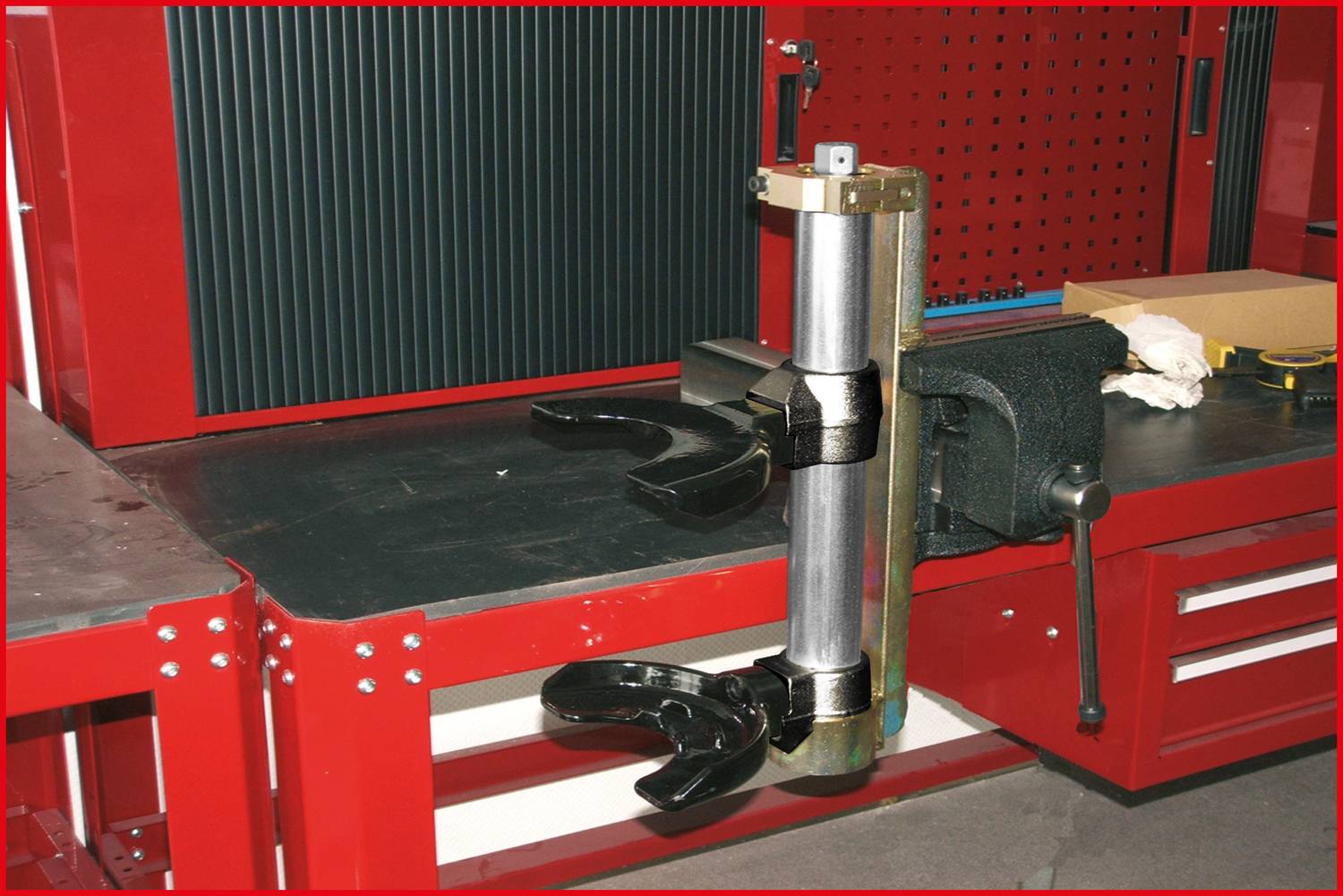 A metal device with two gripping arms is mounted on a workbench in a workshop, surrounded by red cabinets and tools.