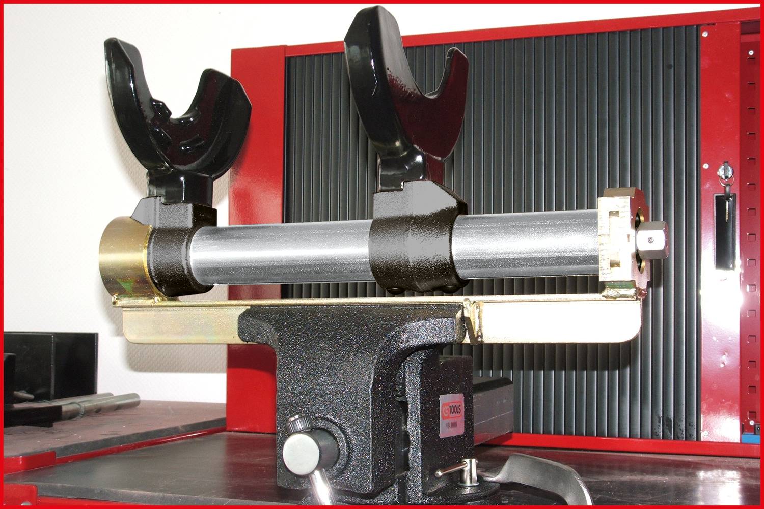 Metal device with two black clamps attached to a horizontal pipe, resting on a sturdy base against a ribbed background.