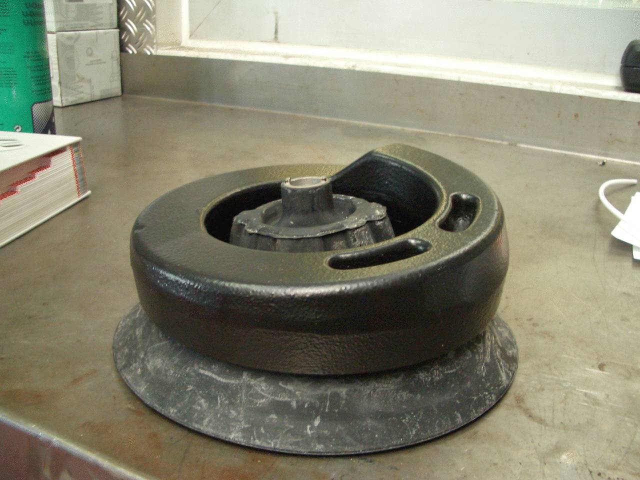 A heavy vehicle component, likely a brake drum, is resting on a workshop table. Tools and a book are visible in the background.