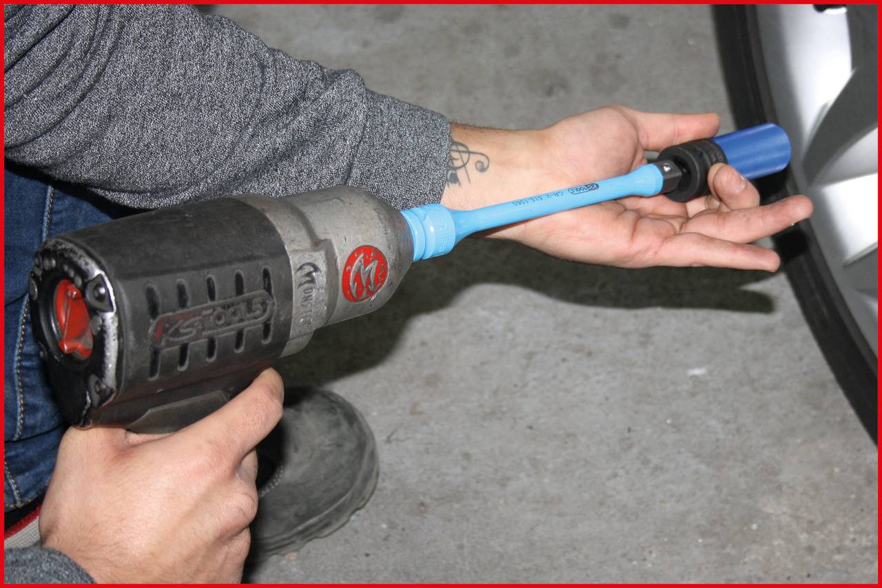 A person is holding an electric hammer drill with a blue extension, pointing it at a car tyre.