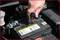 A hand is checking the fluid level of a car battery with a screwdriver under the bonnet. Battery warning signs are visible.