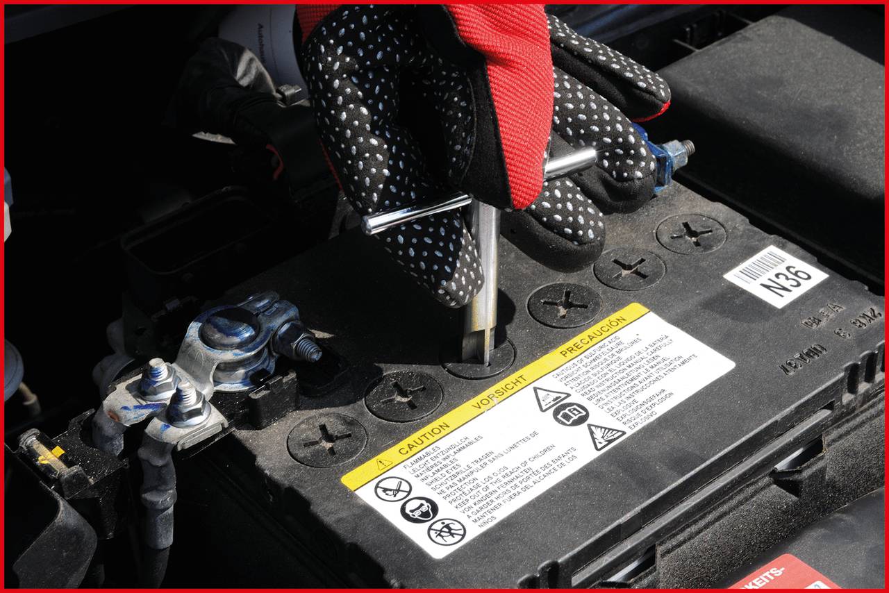 A person wearing red and black gloves is repairing a car battery. The battery has warning stickers and is being worked on with a spanner.