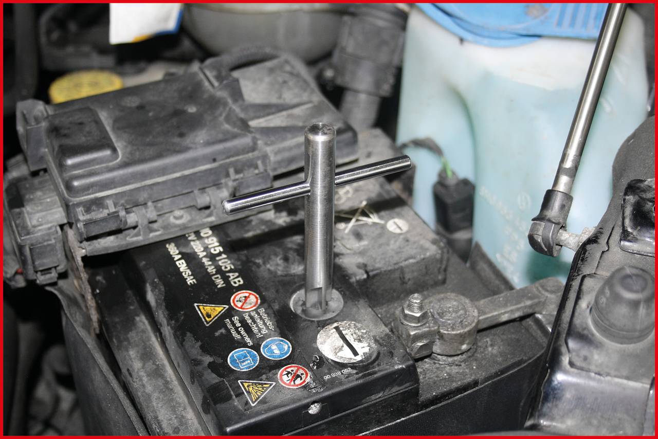 Car battery being secured with tools. A windscreen washer fluid reservoir in the background. Environmental and safety information on the battery.