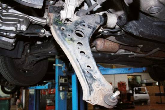 A worn suspension control arm beneath a vehicle on a car lift in a workshop, identifiable by rust marks and wear.