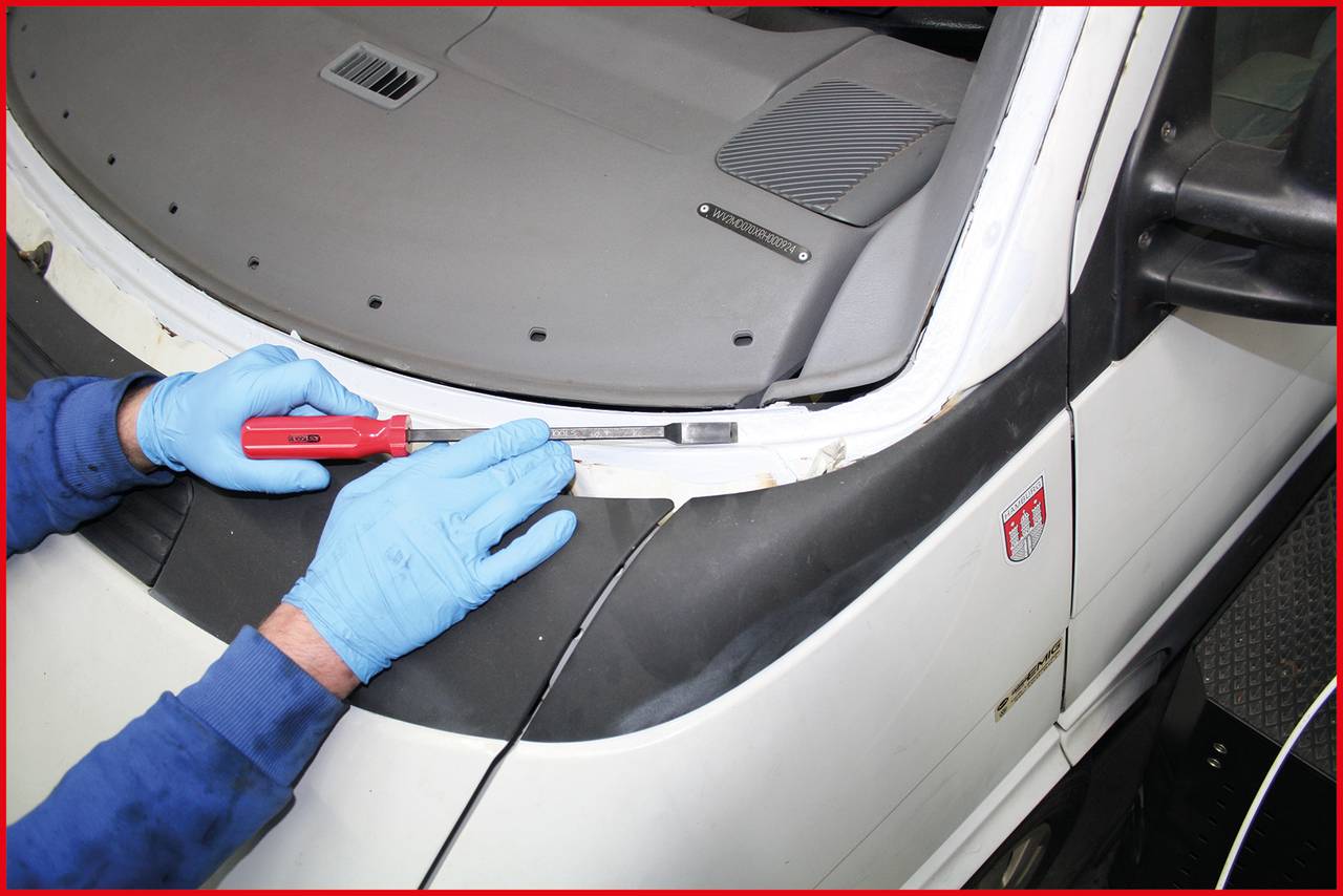 A person wearing blue gloves is using a red cutting tool to remove the sealant from a vehicle's windscreen.