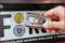 A hand holds a craft knife over a vehicle registration plate with an inspection sticker and label from the Offenbach district, Hesse.