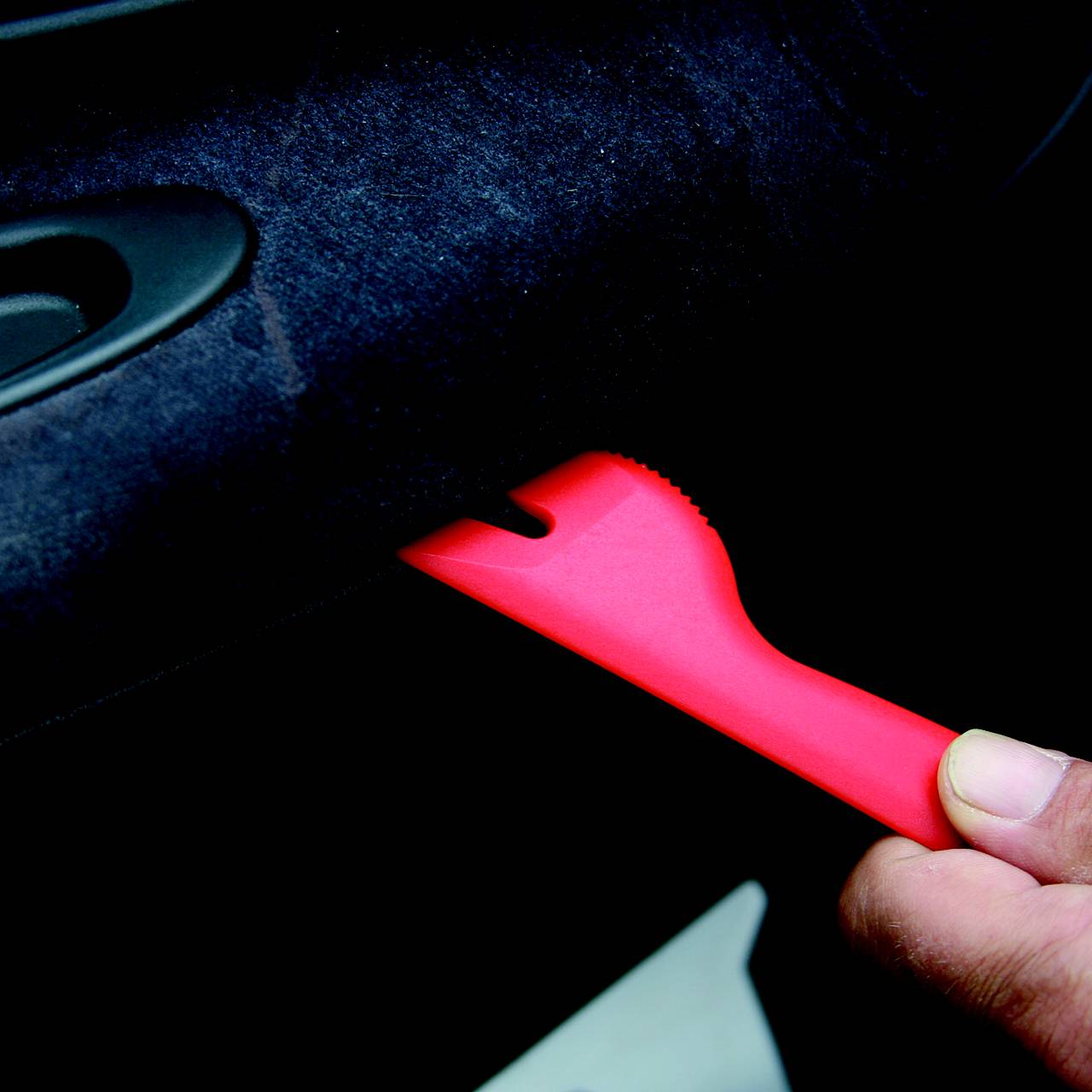 'Hands are holding a red plastic tool near a car door handle.'