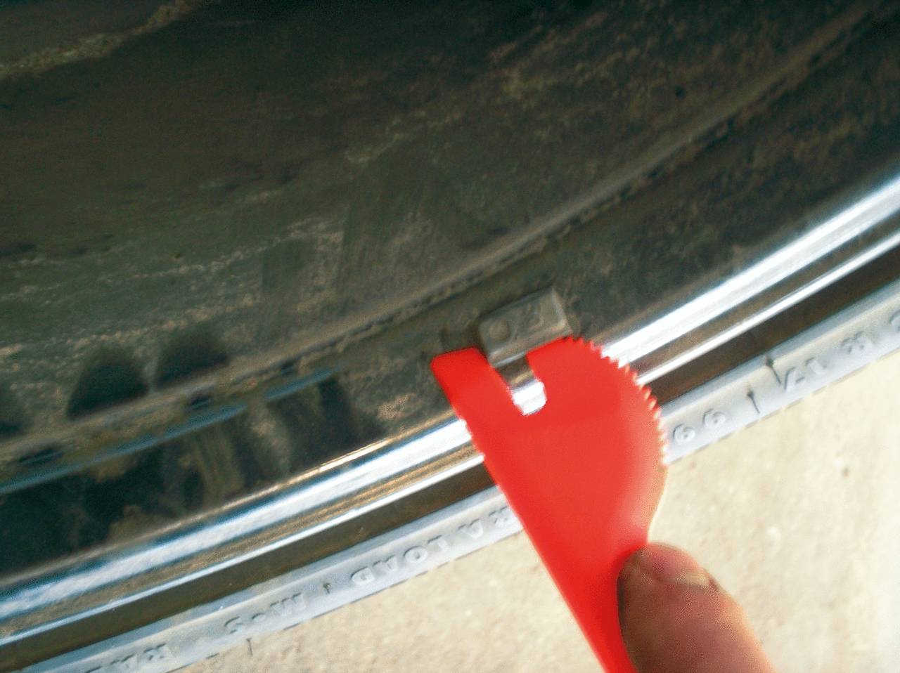 Close-up of a red plastic tool tip removing a tyre weight from a car tyre. The tool has a semi-circular serrated edge.