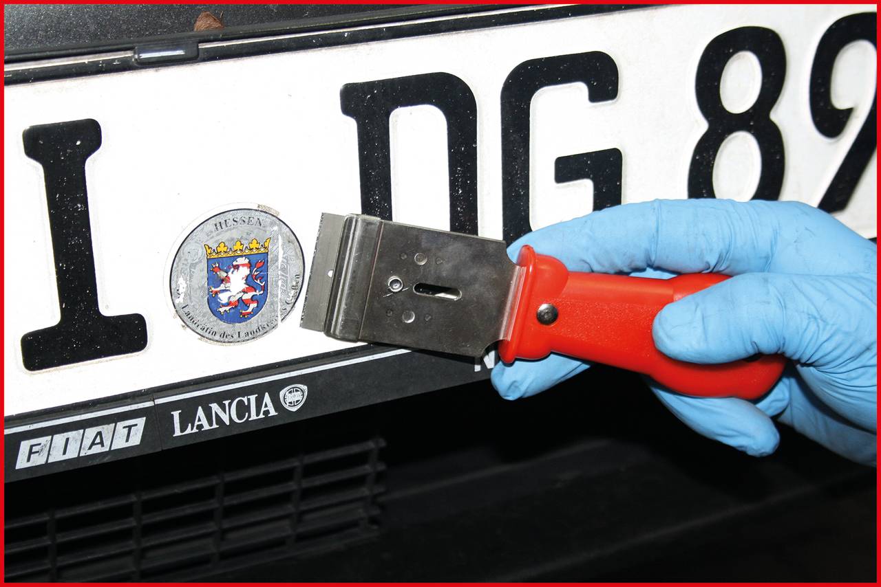 A person wearing a blue glove is removing a sticker from a vehicle number plate using a scraper.