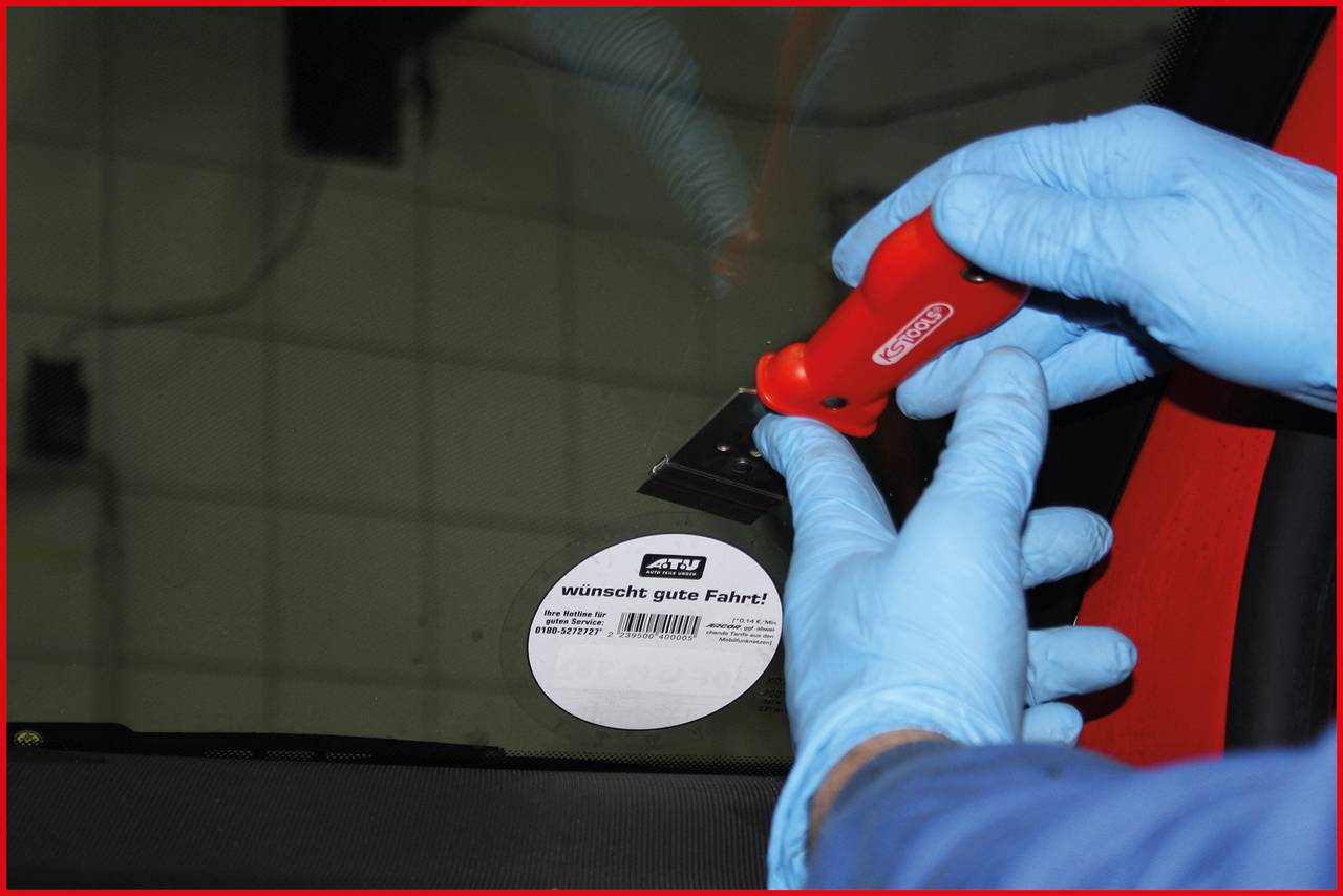 A person is removing a sticker from a car windscreen using a red tool. The sticker reads 'wishes good journey!'