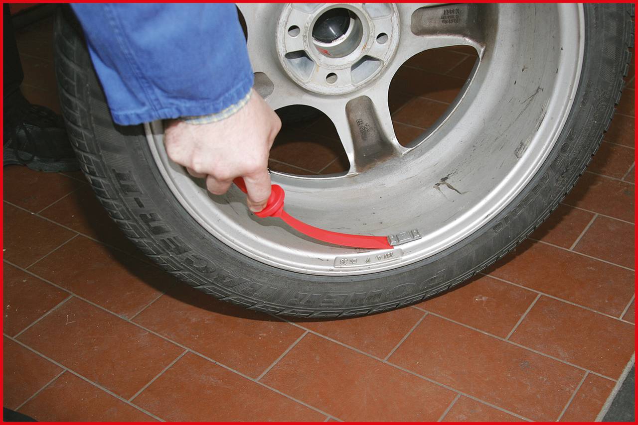A person is checking the air pressure of a car tyre using a red tool on a terracotta-coloured tiled floor.
