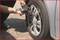 A person is repairing or checking a car tyre with a tool. The tyre is mounted on a grey-silver car.