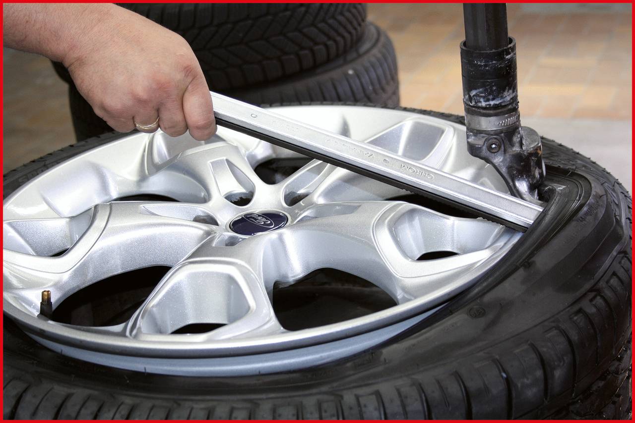 A hand is mounting a car tyre onto an alloy wheel using a tool, with stacked tyres in the background.