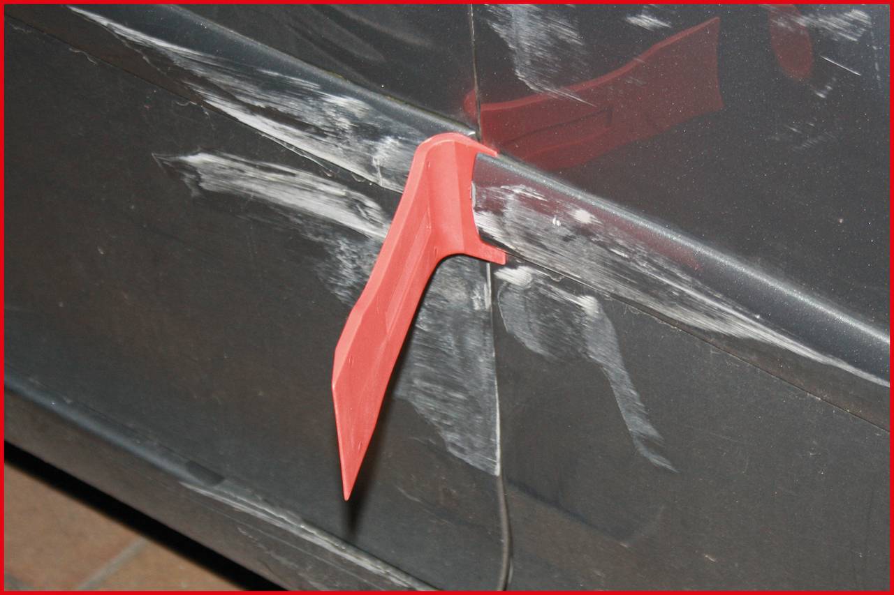 A red plastic cutting tool is stuck in a black, scratched car door panel, apparently used to remove or repair something.