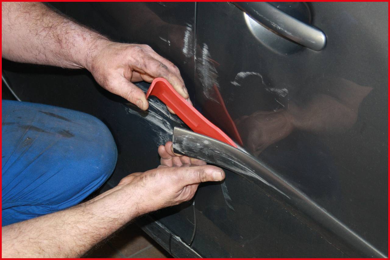 A car mechanic repairs the paintwork on the car door using a filler tool. His right hand holds the door steady, while his left hand uses the tool.