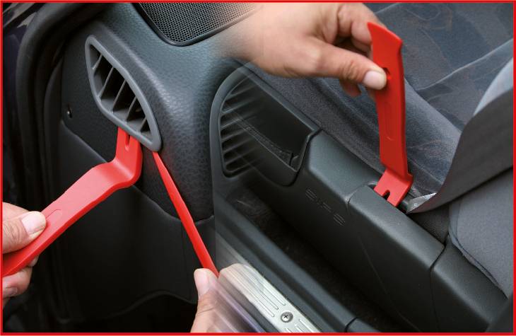 Two red plastic tools are being used to remove parts of a car. One is releasing a ventilation opening, the other a trim panel.